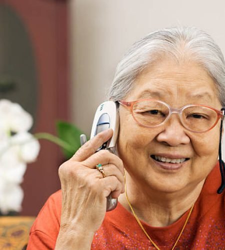 An Asian senior woman, Chinese grandmother on mobile cell telephone, retiree listening, talking and smiling as she communicates with friend and family from her home. Orchid flowers and Chinese furniture are in soft focus in the residential interior.
